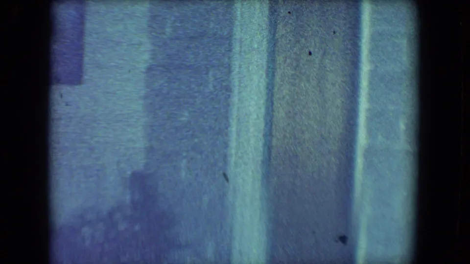 1961 Salton Sea home-movie frame showing a dim living room edge with vertical curtains or wall panels and blue-green film tint.