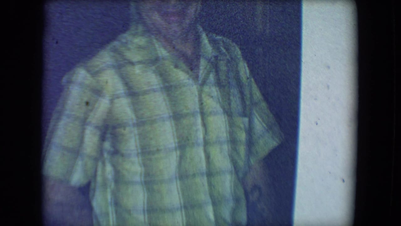 Man in a green plaid shirt stands in a dim room with his face mostly out of frame.