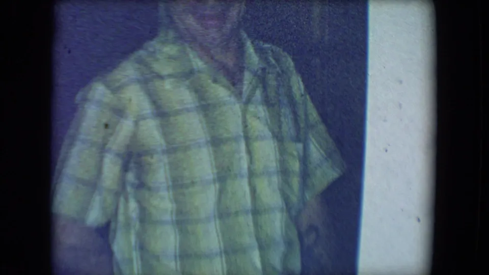 1961 8mm home-movie frame from Salton Sea, California showing a man in a green plaid shirt in a dim private interior.