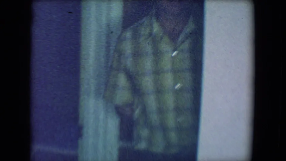 1961 Salton Sea home-movie frame with a man in a green plaid shirt partly turned from the camera inside a dim room.