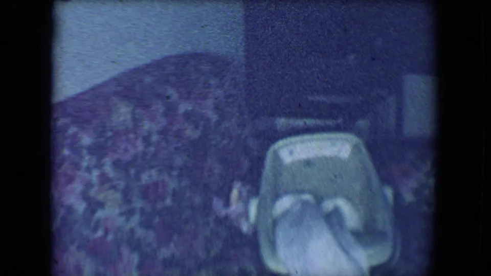 A dim 1961 home-movie frame shows a baby carrier near patterned fabric and dark furniture or shadowed interior space.