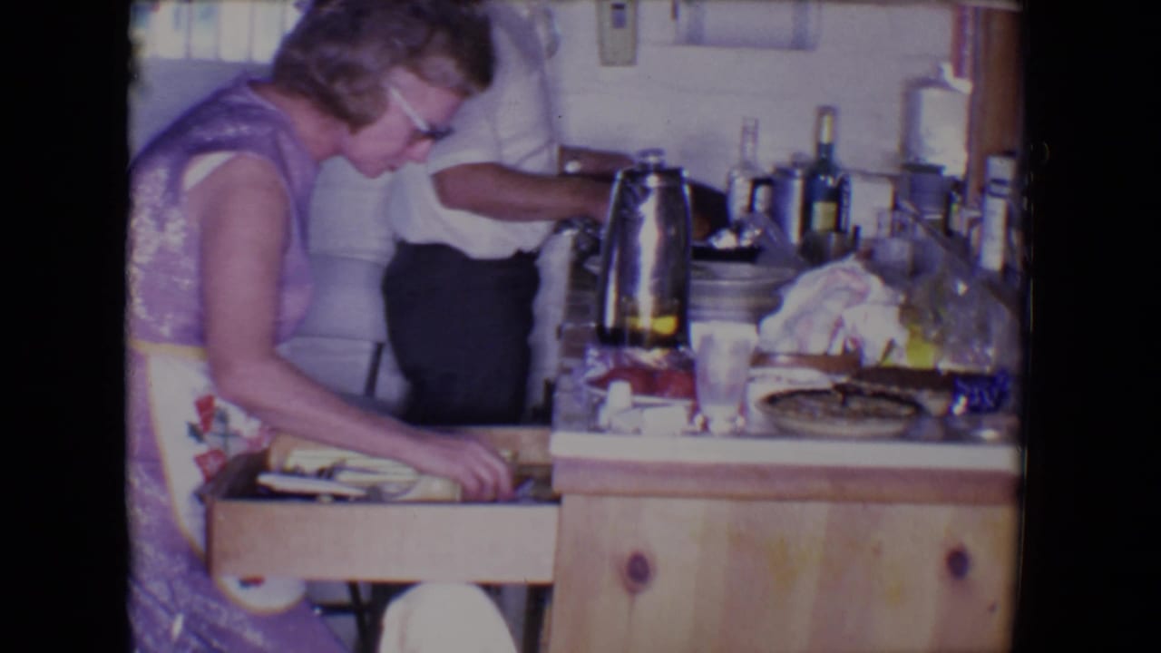 In a 1961 Salton Sea home kitchen, a woman in glasses searches a wooden drawer as a man stands nearby behind a cluttered counter.