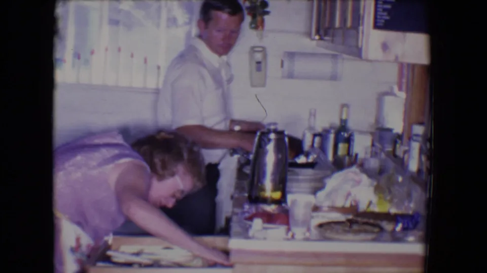 The woman leans deeply into the drawer area while the man remains near the counter in the 1961 Salton Sea kitchen scene.