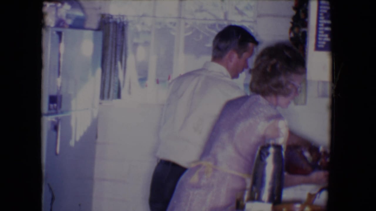 The woman reaches near the counter as the man assists in the 1961 Salton Sea kitchen, with refrigerator and curtains behind them.