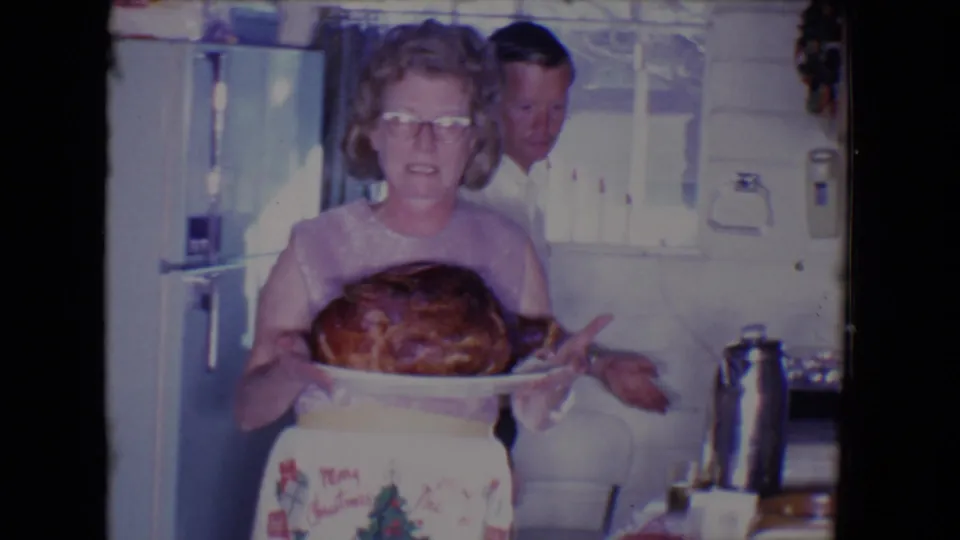 A woman in glasses and an apron presents a roasted turkey on a platter in the 1961 Salton Sea home kitchen scene.