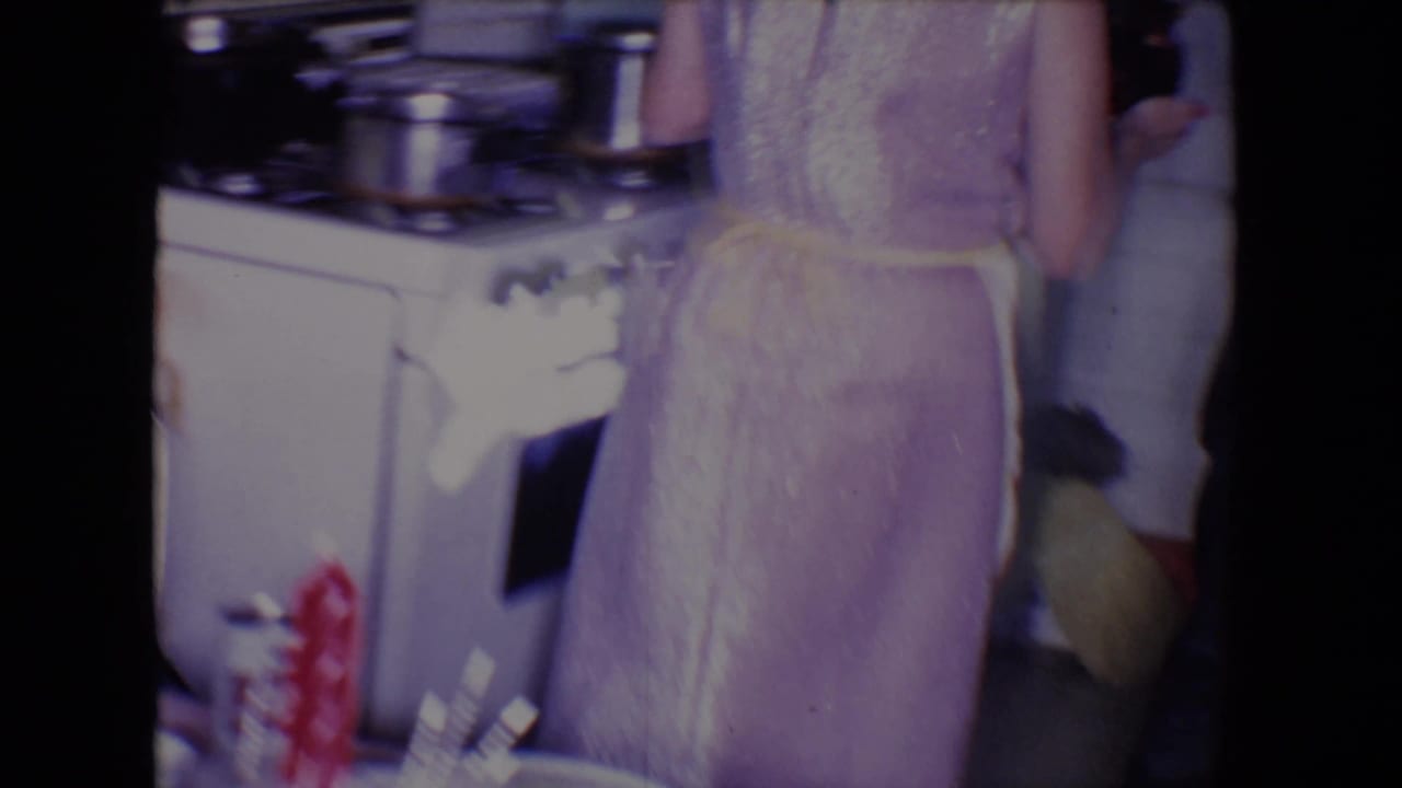 Woman in a purple dress stands near a stove with kitchenware visible around her.