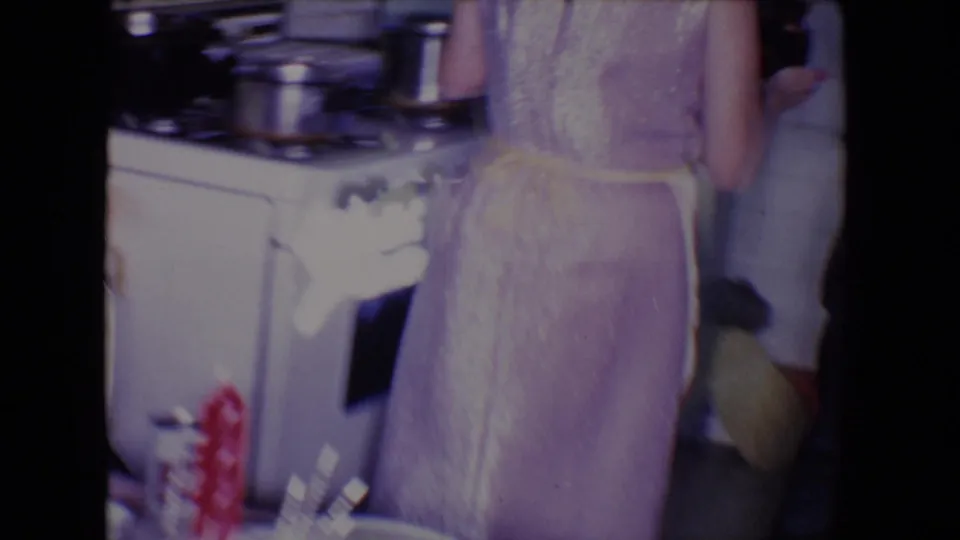 A woman in a purple dress is seen from the side near a gas range in the 1961 Salton Sea kitchen scene.