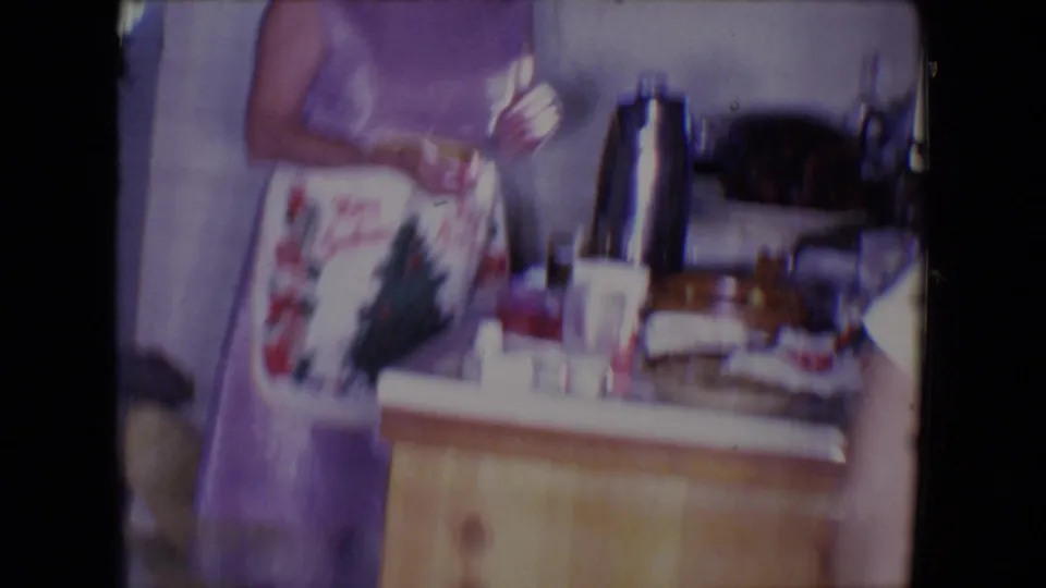 The woman stands beside a cluttered kitchen counter with drinkware, a metal pot, and holiday apron details visible.