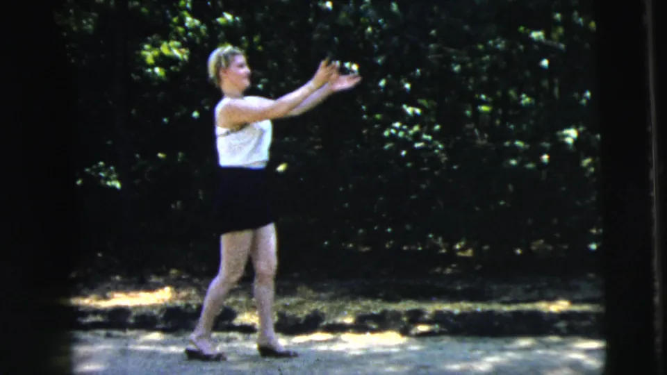 In the supplied 1962 Camden context, a young woman stands in a shaded park area reaching toward a small white ball.