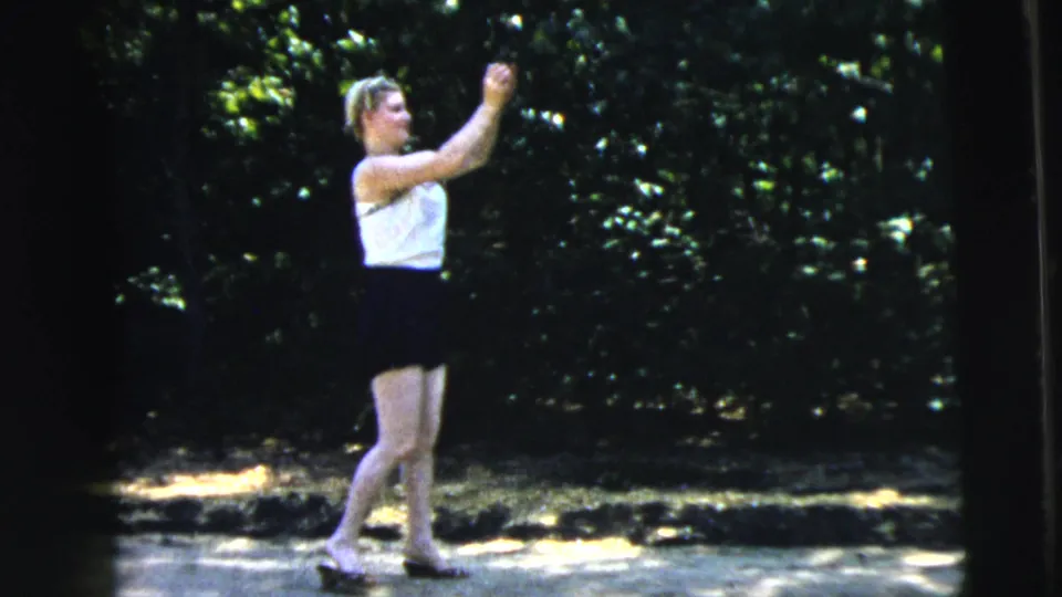 A young woman in a white top and dark lower garment raises her arm during a ball toss in the 1962 Camden home-movie scene.