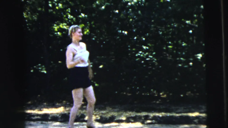 The woman pauses or steps forward in the Camden park setting, with dense trees and dappled light behind her.