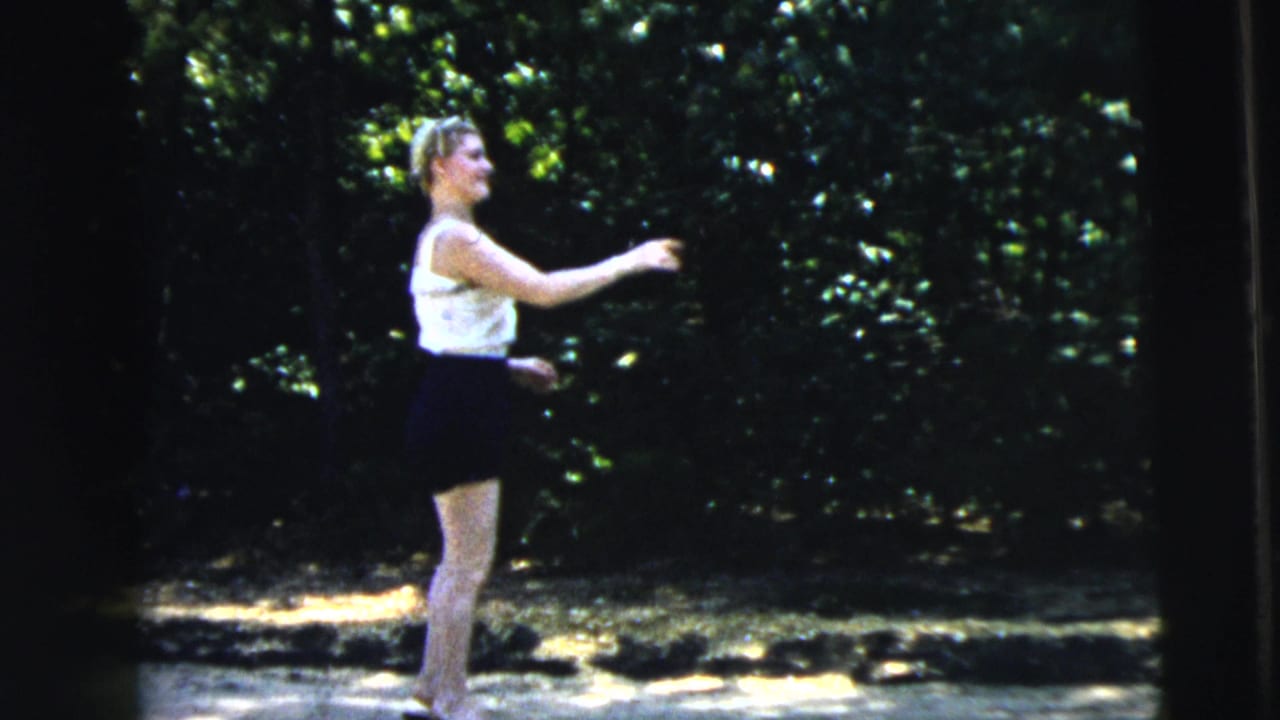 A teenage girl in a white top and dark skirt reaches forward on a sunlit Camden roadside in 1962 8mm home-movie footage.