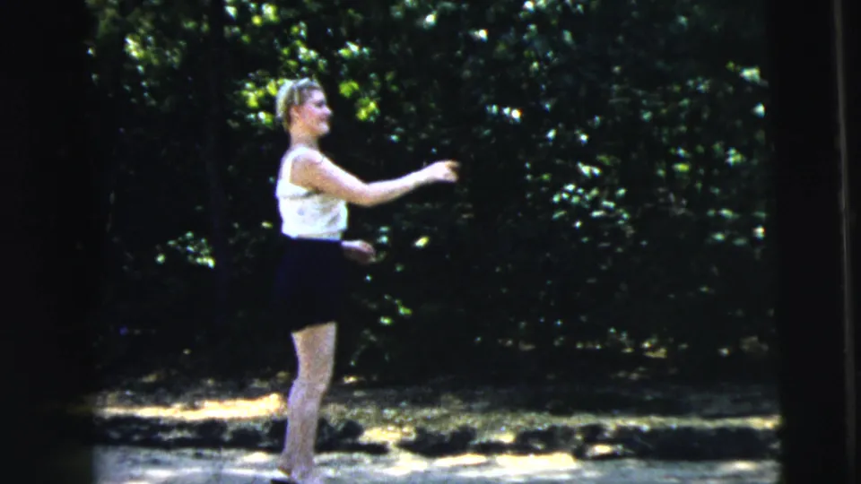 A teenage girl in a white top and dark skirt reaches forward on a sunlit Camden roadside in 1962 8mm home-movie footage.