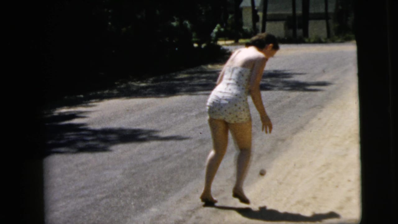 Woman in a light polka-dot swimsuit bends toward a small ball on a sunlit road.