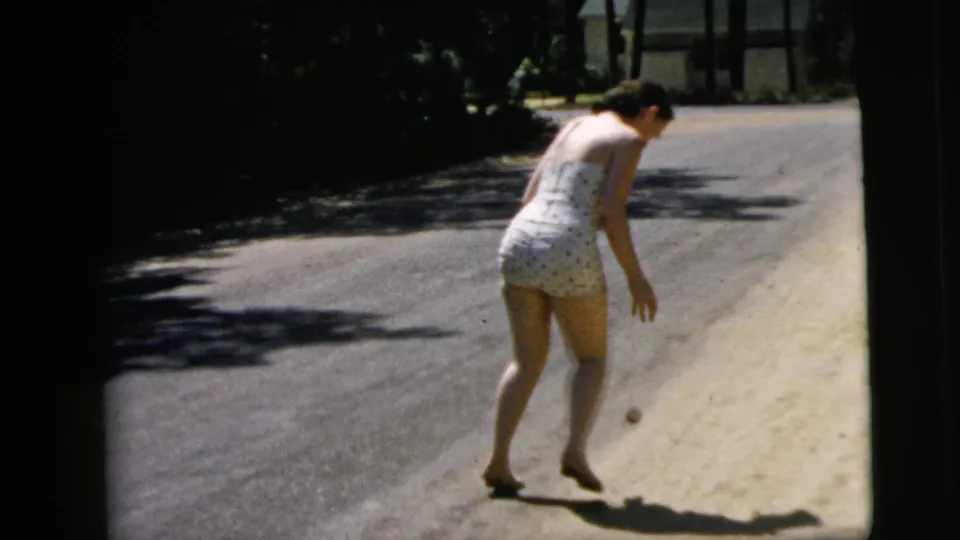 In Camden, New Jersey in 1962, a woman in a polka-dot swimsuit bends toward a small ball near the edge of a sunny road.