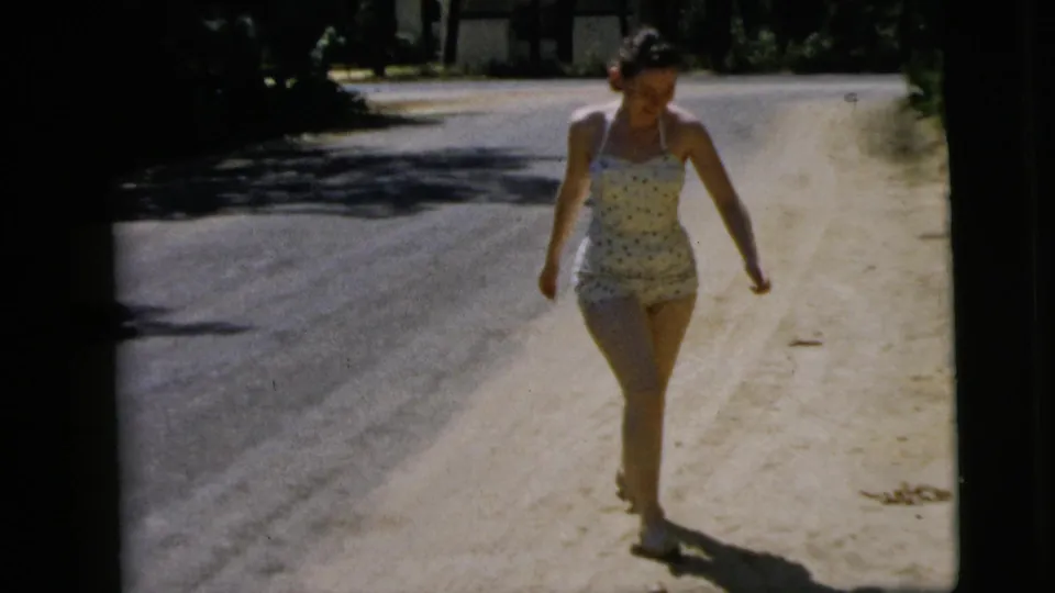 A woman in a light patterned swimsuit walks toward the camera on a quiet Camden road in the 1962 home-movie footage.