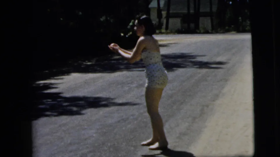 The woman stands side-on on the road with her hands raised, appearing ready to catch or handle the small ball in 1962 Camden footage.