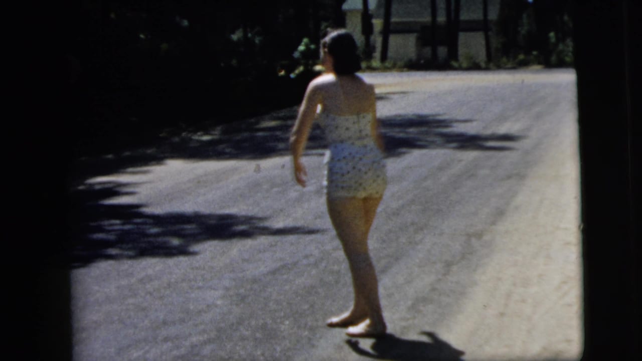 In 1962 Camden, New Jersey home-movie footage, a barefoot woman in a patterned swimsuit stands on a sun-dappled road.