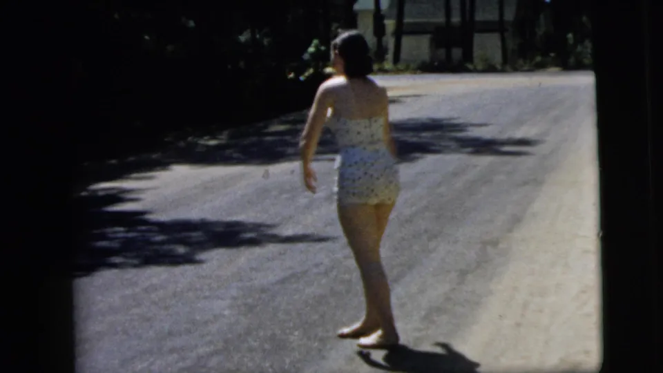 In 1962 Camden, New Jersey home-movie footage, a barefoot woman in a patterned swimsuit stands on a sun-dappled road.