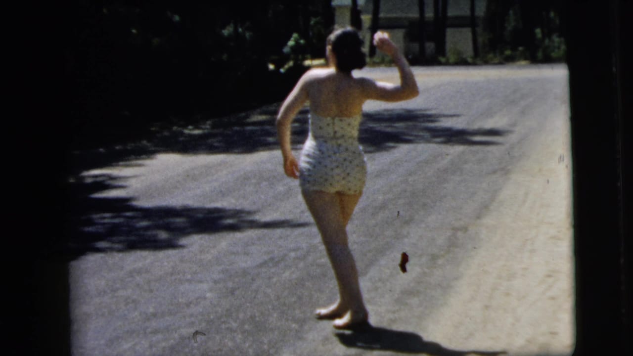 Woman in swimsuit raises one arm while standing barefoot on the road.