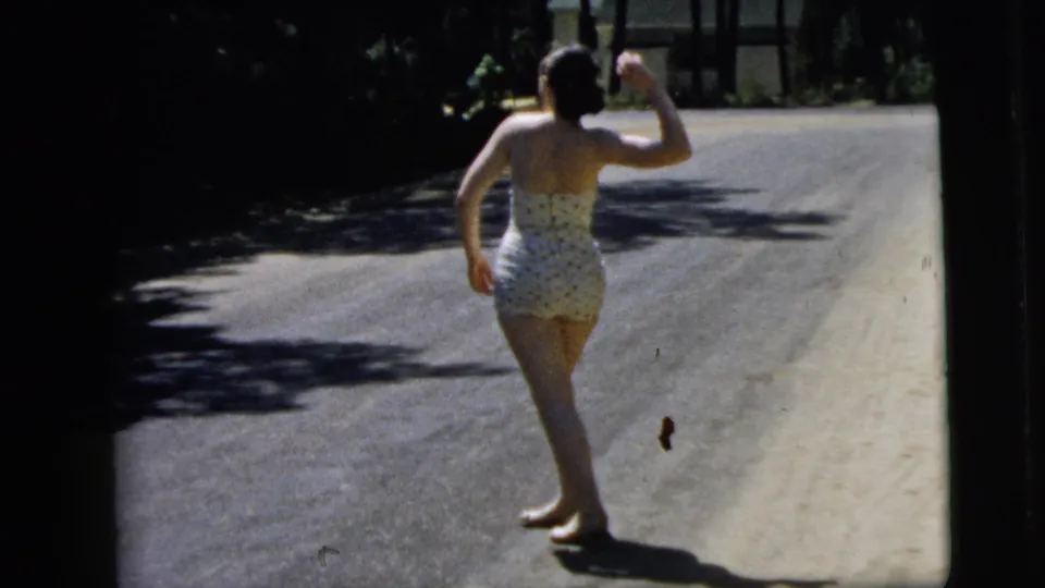The woman raises an arm as if preparing to throw a small object on a shaded Camden road in 1962 footage.