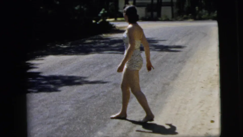 The barefoot woman walks across the road after the gesture, her shadow visible in the 1962 Camden home-movie frame.
