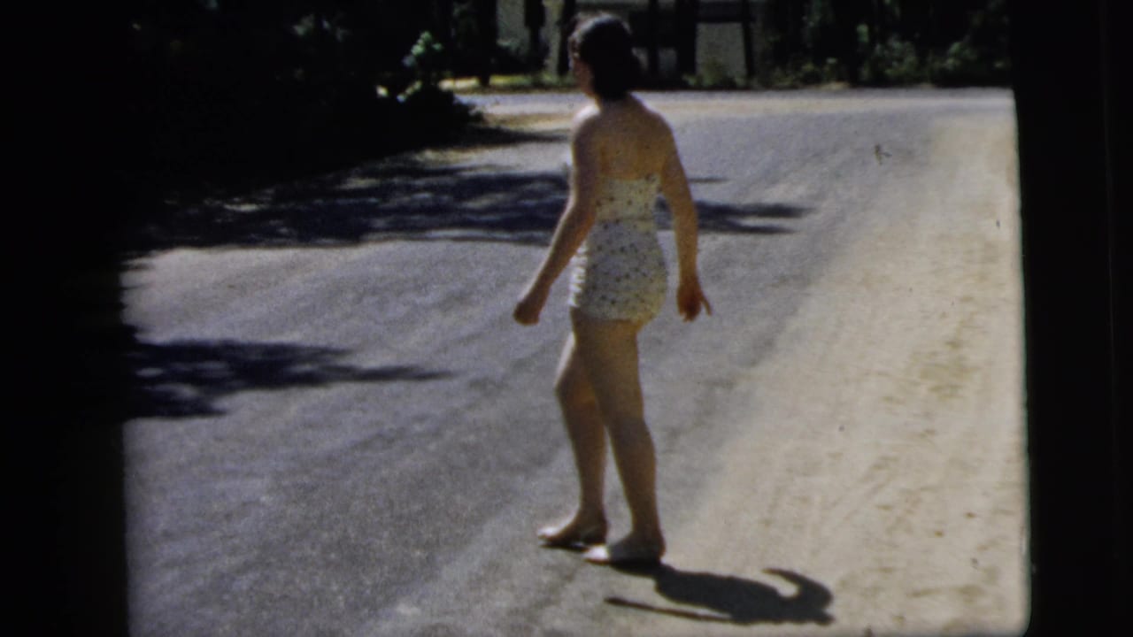 Woman in a patterned one-piece swimsuit walking barefoot on a sunny road shoulder.