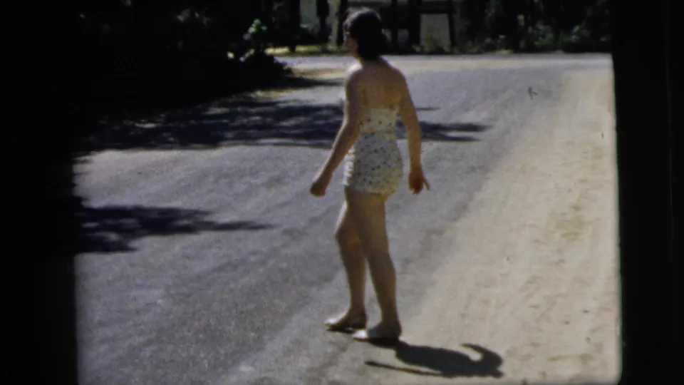In 1962 Camden, New Jersey home-movie footage, a woman in a white patterned swimsuit walks barefoot beside a sunlit road.