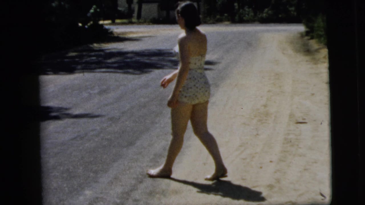 The woman continues away from the camera on a sunlit Camden road, with tree shadows crossing the asphalt and shoulder.