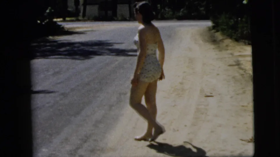 A side-rear view shows the barefoot woman turning slightly as she walks along the bright road shoulder in Camden, New Jersey.