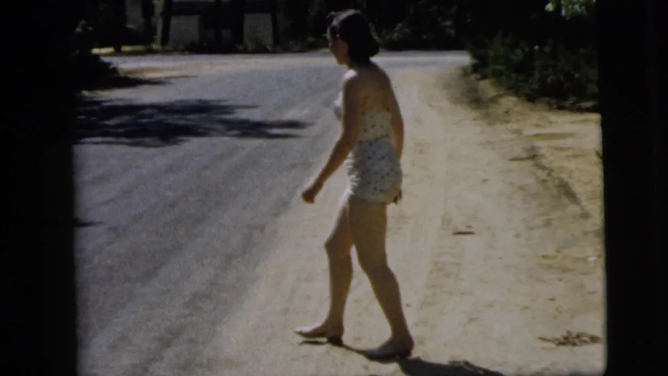 The final frame shows the woman in a patterned swimsuit walking beside the paved road, with bright shoulder light and foliage beyond.