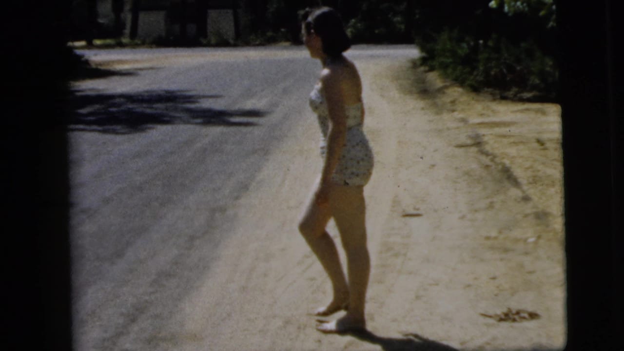 Young woman in a polka-dot swimsuit stands barefoot beside a sandy roadside.