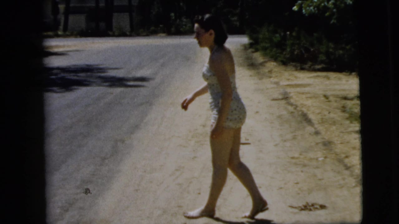 The young woman continues walking barefoot along a sandy roadside in Camden, New Jersey, with trees and road behind her.