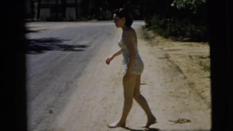 The young woman continues walking barefoot along a sandy roadside in Camden, New Jersey, with trees and road behind her.