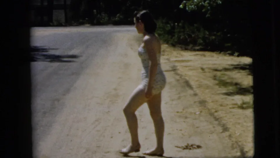 A 1962 home-movie frame shows the woman near the sandy edge of the road, her dark bob and polka-dot swimsuit visible.