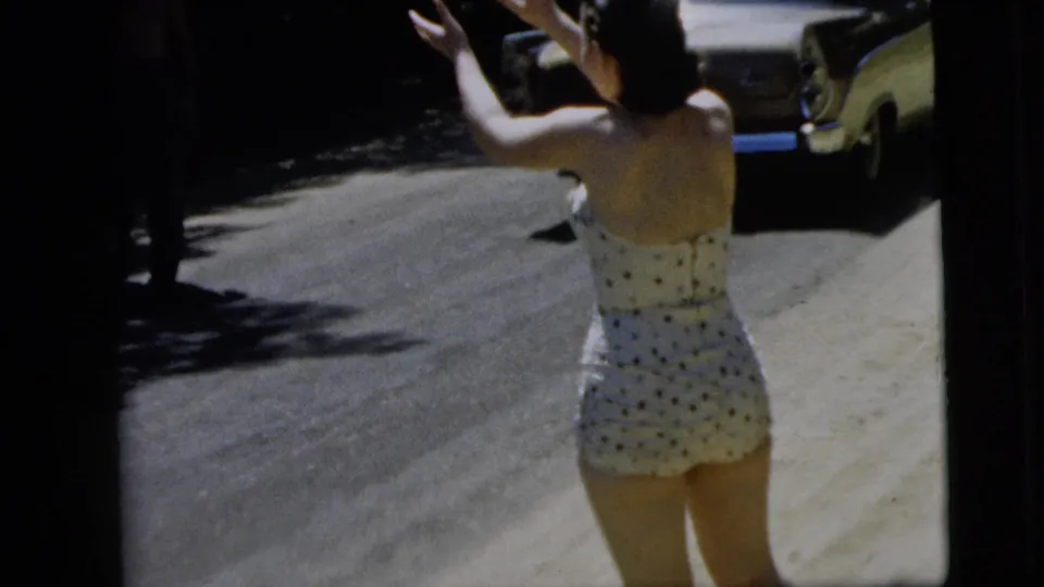 The woman turns away from the camera and raises her arms near the road as a vintage car is visible in the background.