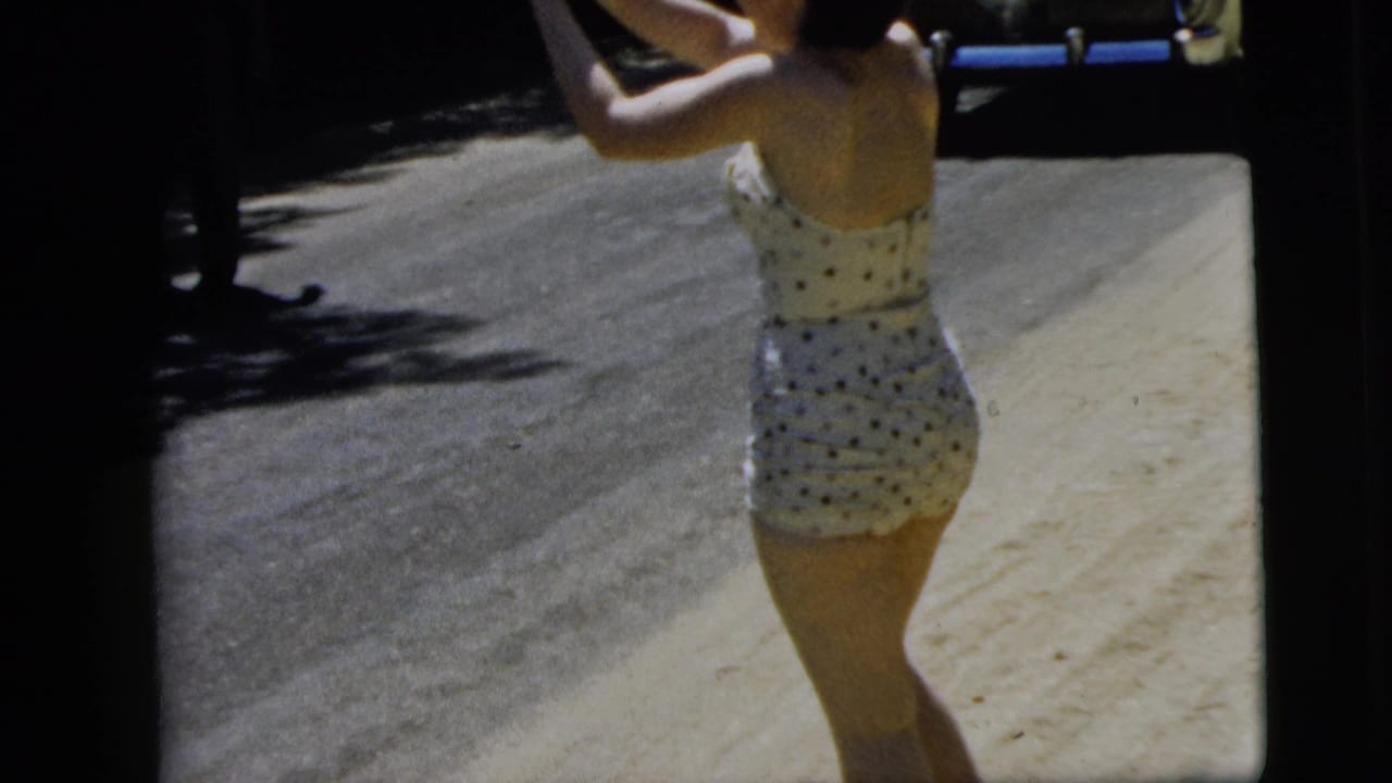 In Camden, New Jersey in 1962, a young woman in a white polka-dot swimsuit stands on a sunlit residential road with her arms raised.