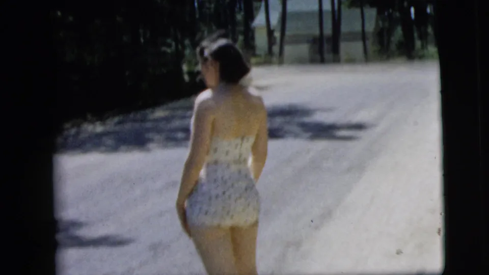 The woman in a white polka-dot swimsuit walks away down a quiet Camden road, with trees and a distant house visible in the background.