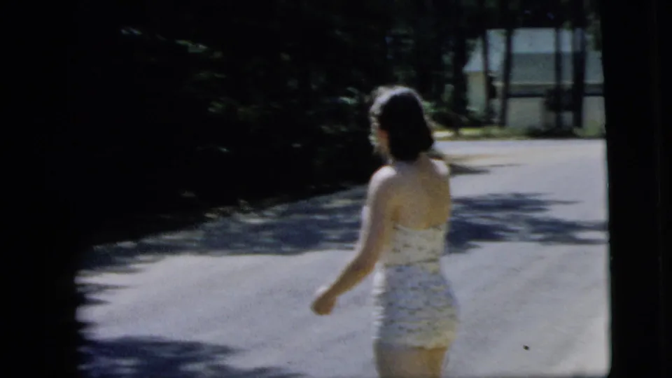 In metadata-supported 1962 Camden, New Jersey footage, a woman in a white polka-dot swimsuit walks across a bright residential street.