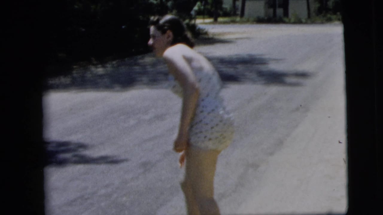 The woman bends slightly forward on a sunlit Camden street, with tree shade and a pale paved road behind her.