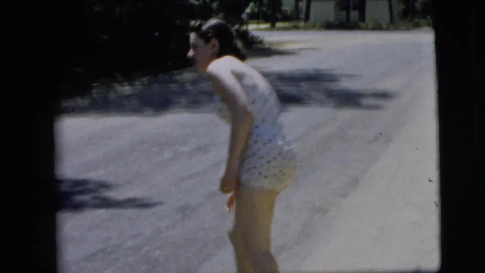 The woman bends slightly forward on a sunlit Camden street, with tree shade and a pale paved road behind her.