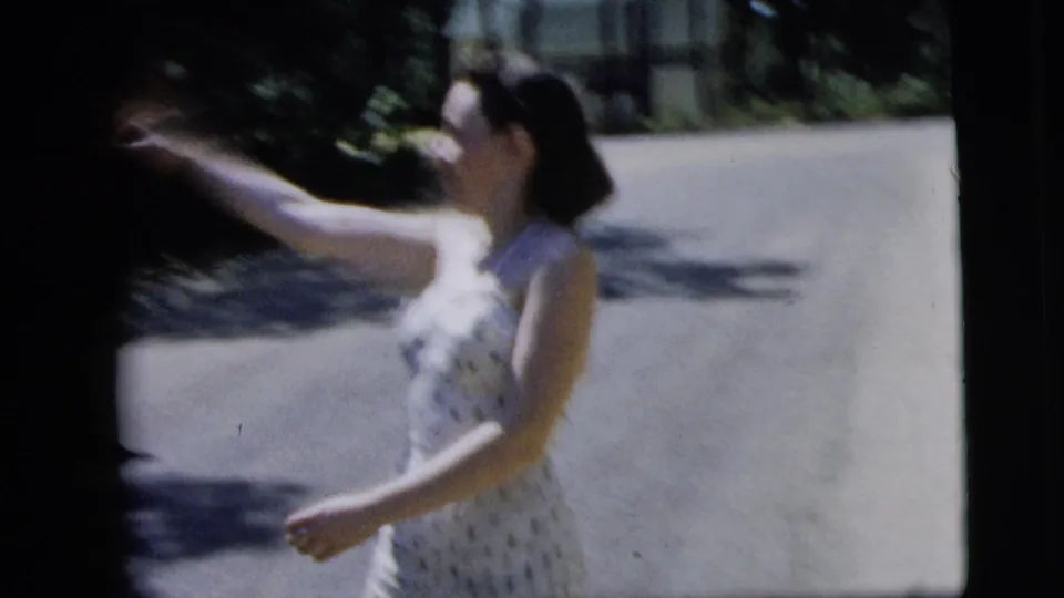 A woman in a polka-dot swimsuit raises an arm in a playful gesture on the bright residential street, as captured in soft 8mm focus.