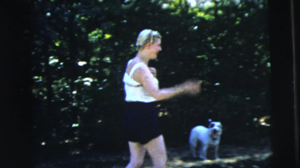 In a wooded area, the woman appears in a sleeveless top and dark shorts near a small light-colored dog standing by the trees.