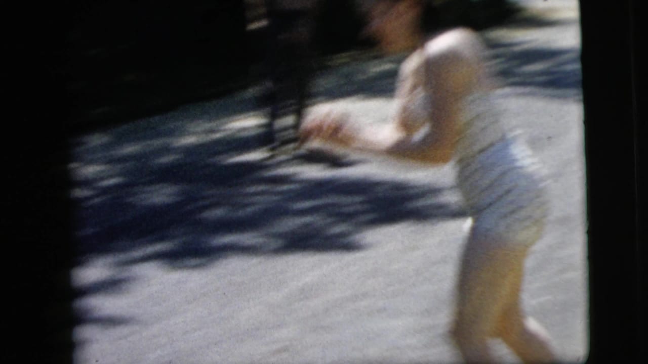 Young woman in a polka-dot swimsuit moves across a bright residential street in the 1962 Camden, New Jersey home-movie footage.
