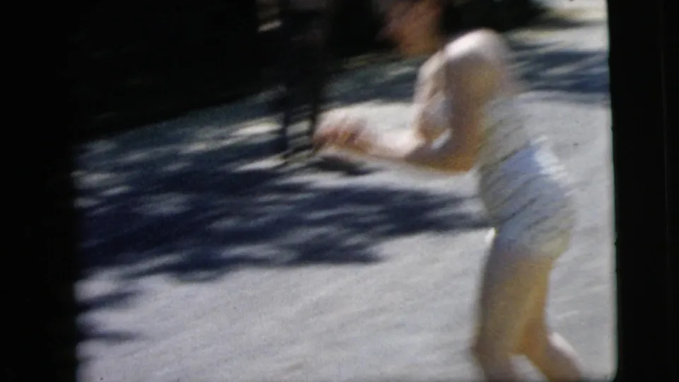 Young woman in a polka-dot swimsuit moves across a bright residential street in the 1962 Camden, New Jersey home-movie footage.