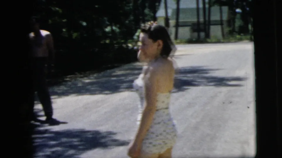 A woman in a polka-dot swimsuit stands on the paved street while a shirtless man appears at the shaded edge of frame.