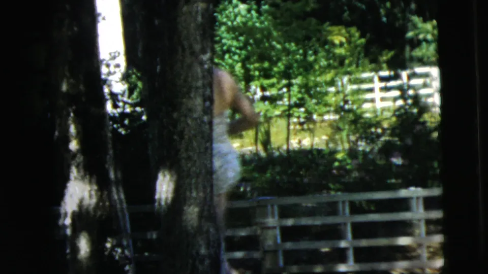 The woman in the swimsuit is partly obscured by tree trunks beside a white fence and greenery in the Camden home-movie scene.