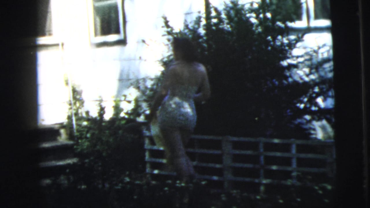Woman walks beside shrubs toward the house, with bright siding and a low fence behind her.