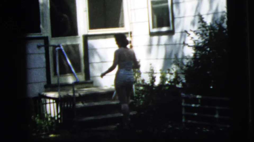 The woman reaches the small porch area of the Camden residence, framed by steps, a doorway, windows, and a metal rail.