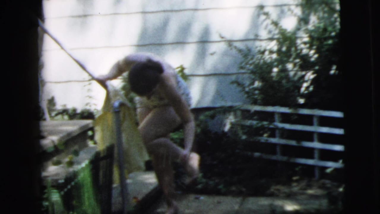 In Camden, New Jersey, 1962 home-movie footage, a young woman in a swimsuit bends near a garden gate with a yellow towel.